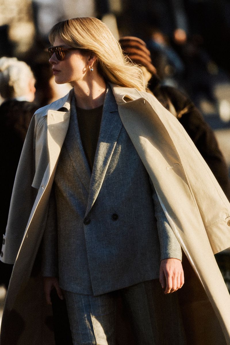 Woman in a grey tailored suit and draped beige trench coat wearing sunglasses on a sunny city street.