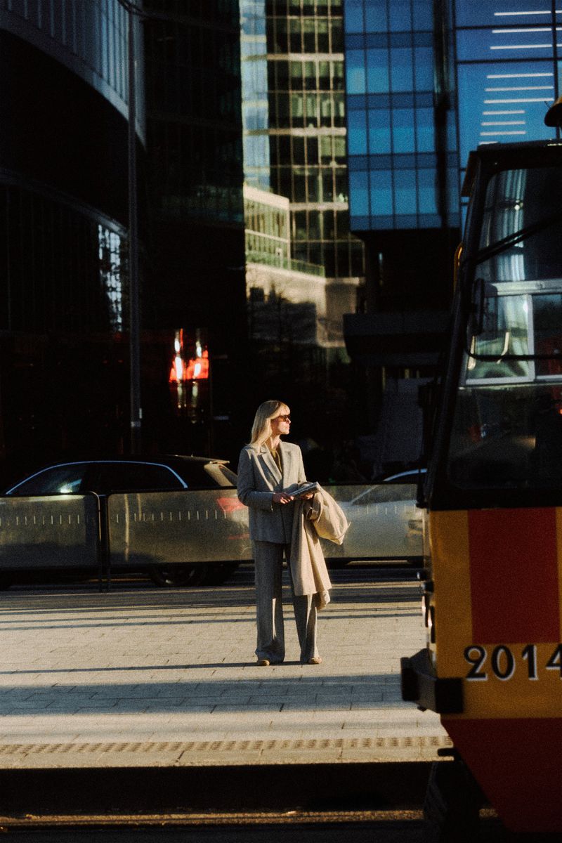 Woman in a grey pantsuit standing on a paved city plaza with modern skyscrapers and cars in the background