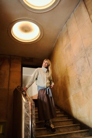 Model standing on a stone staircase under circular ceiling lights wearing a light beige jacket and a long brown tiered skirt.