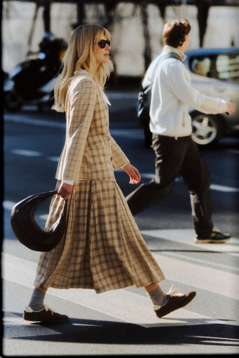 Woman in plaid blazer and pleated midi skirt crossing a city street carrying a dark handbag, captured with a grainy film aesthetic.