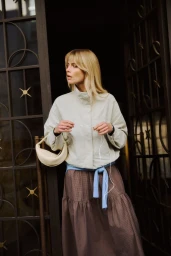 Blonde woman in a pale grey jacket and tiered brown skirt holding wired earphones in front of a decorative iron doorway.