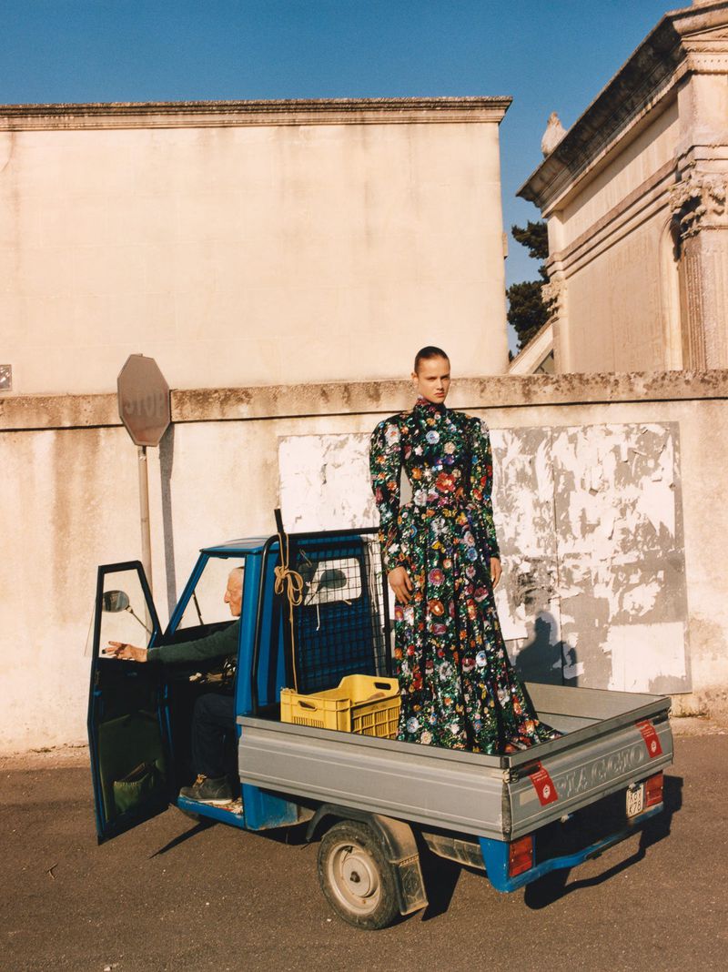 Model standing on the back of a blue truck wearing a dark floral long sleeve dress outdoors by a wall