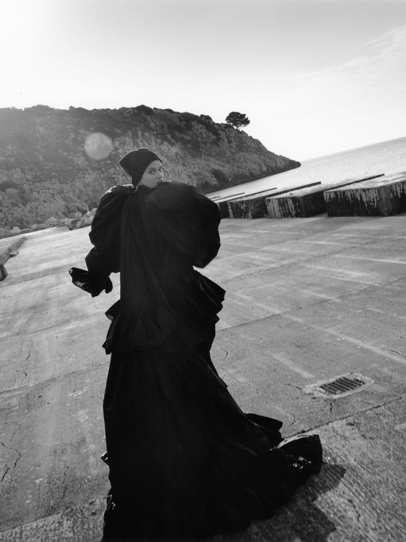 Model Noor Khan wearing a voluminous black gown and beanie walking along a coastal concrete pier in a black and white editorial shot.