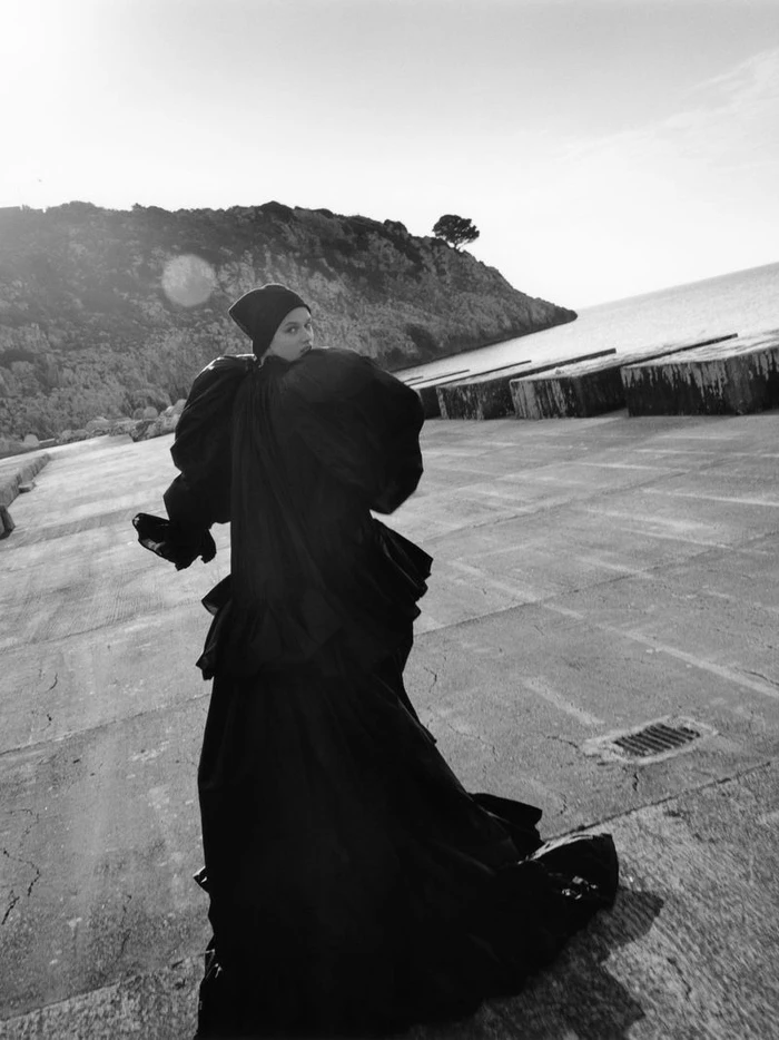 Model Noor Khan wearing a voluminous black gown and beanie walking along a coastal concrete pier in a black and white editorial shot.