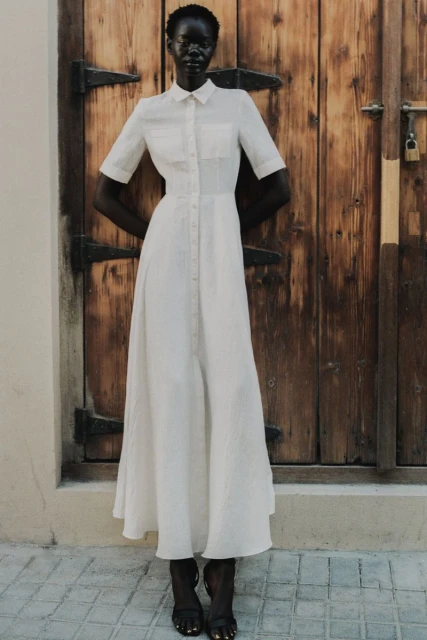 Elegant dark-skinned woman poses in a full-length button-down white dress and black sandals against a rustic wooden door backdrop.