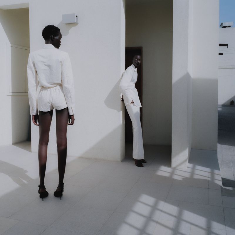 Two women in minimalist white outfits stand against a bright, geometric, all-white architectural backdrop with strong shadows defining the setting.
