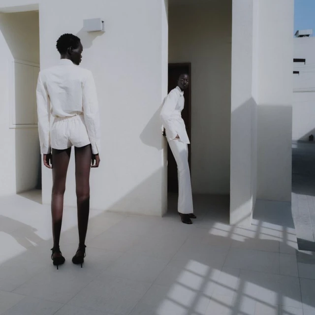 Two women in minimalist white outfits stand against a bright, geometric, all-white architectural backdrop with strong shadows defining the setting.