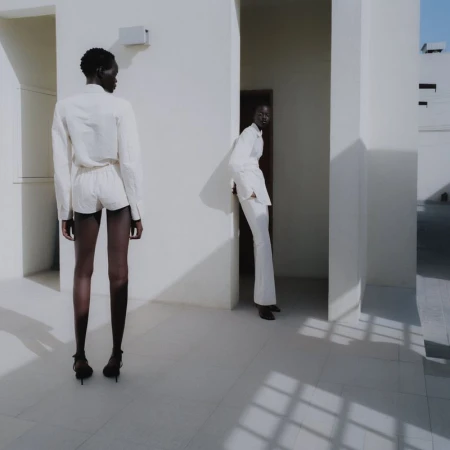 Two women in minimalist white outfits stand against a bright, geometric, all-white architectural backdrop with strong shadows defining the setting.