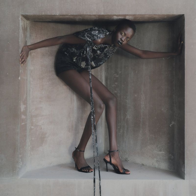 A slender dark-skinned fashion model poses in a concrete niche wearing a printed black top, dark shorts, and high-heeled sandals.