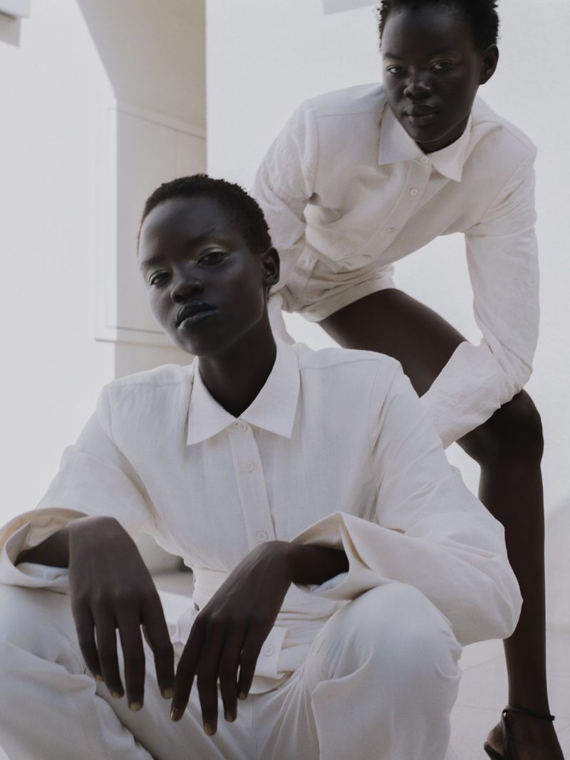 Two dark-skinned adult women pose in a light-filled, minimalist photo studio wearing matching white button-down shirts and trousers.