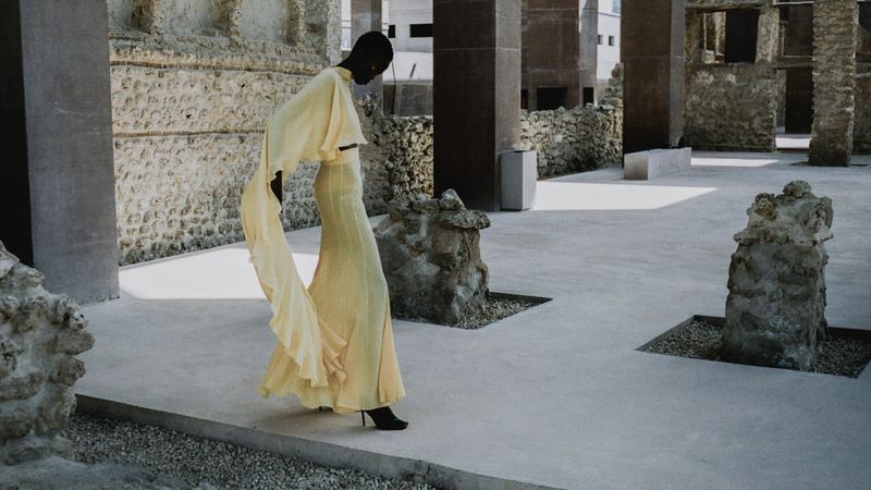 An adult female fashion model in a yellow gown poses in an outdoor courtyard with stone walls and architectural elements, creating an editorial shot.