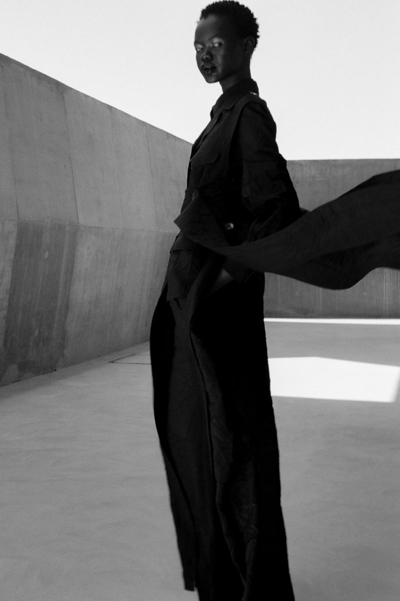 Black and white studio shot of a woman in an all-black outfit standing against a concrete wall, showcasing fashion and style.