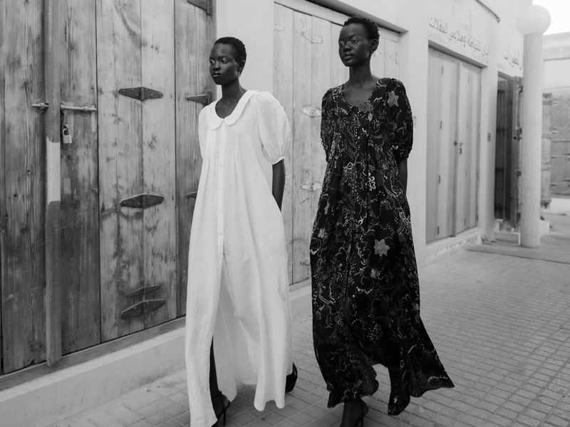 Two women in flowing dresses pose in front of a weathered wooden door and stucco building in a black-and-white outdoor fashion shot.