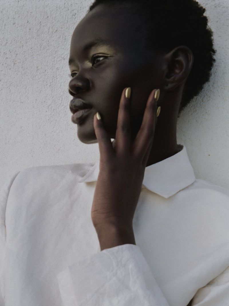 Portrait of an adult female of African descent wearing a white collared shirt with golden makeup and nails against a white wall.