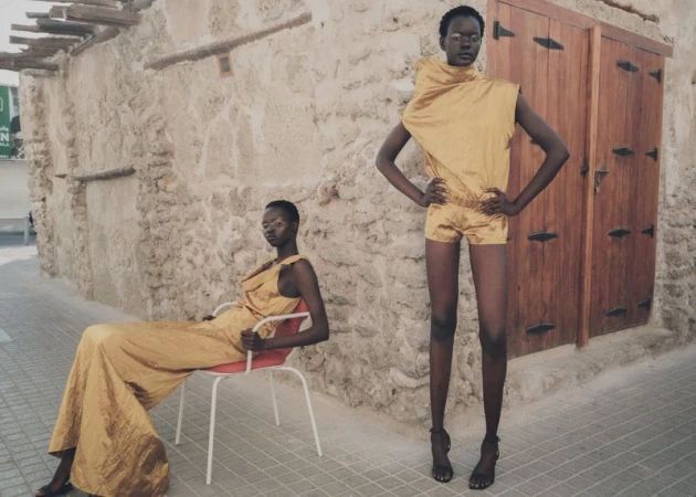 Two Black women in gold outfits pose in front of a stone wall, one seated on a red and white chair and the other standing.
