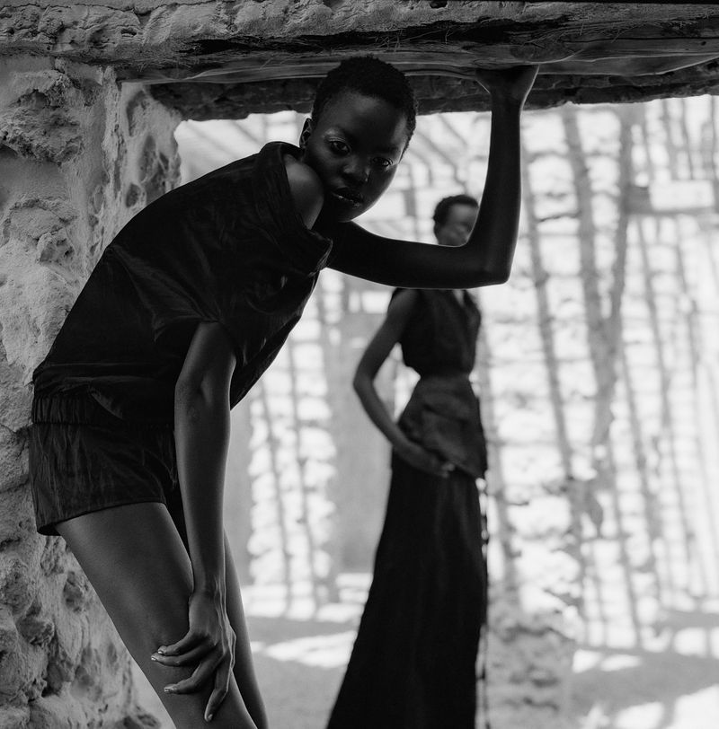 Two Black women pose in black clothing within stone architecture. One leans against stone, the other stands in background, monochrome.