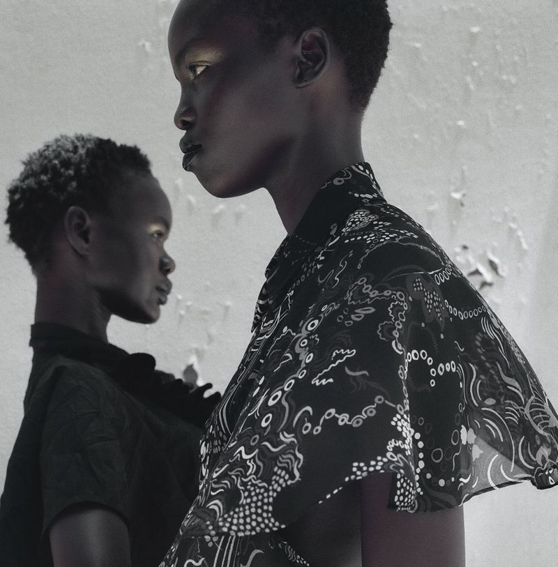 Two Black women in a close-up shot, one in focus with a patterned blouse, the other in soft focus, against a peeling paint wall.