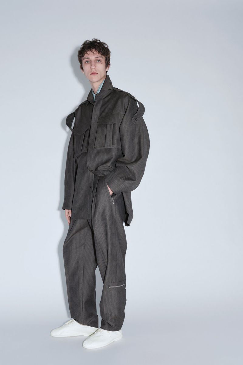 Model Saul Symon posing in an oversized dark grey utility suit and white sneakers against a pale studio seamless background.