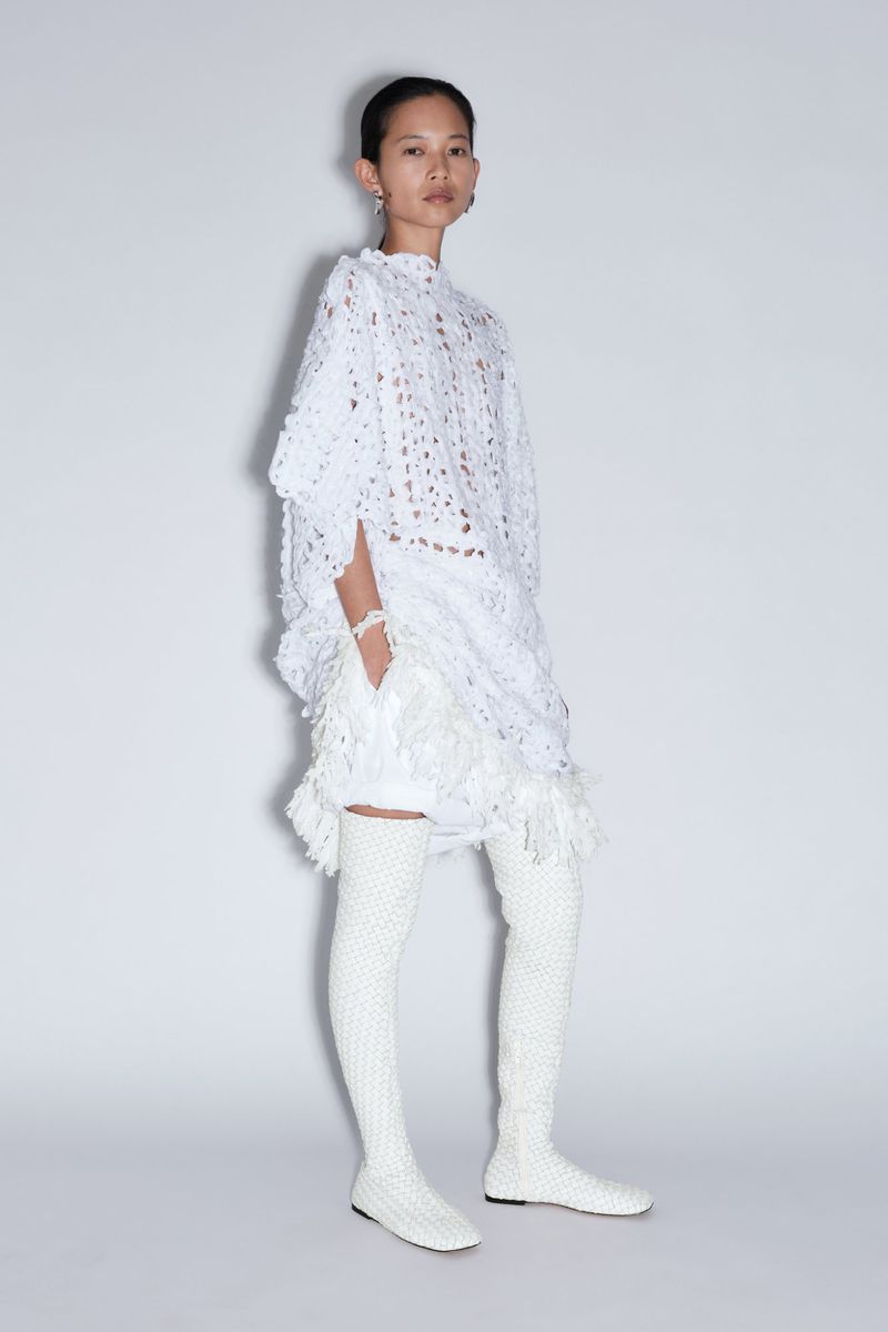 Model posing in an all-white textured lace tunic with fringe trim and matching thigh-high knit boots against a light studio backdrop.