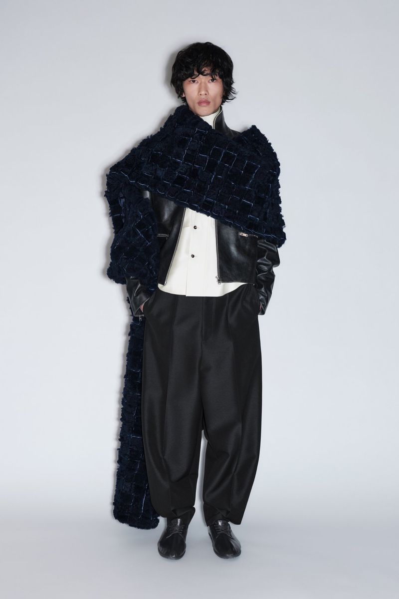 Model wearing an oversized dark textured scarf, black leather jacket, white shirt, and wide-leg black trousers on a grey background.