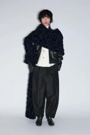 Model wearing an oversized dark textured scarf, black leather jacket, white shirt, and wide-leg black trousers on a grey background.
