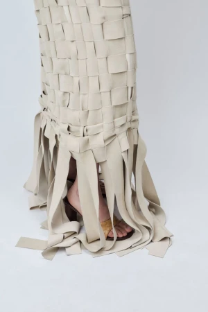 Close-up of a beige woven leather fringe skirt cascading over tan heeled sandals on a seamless white background.