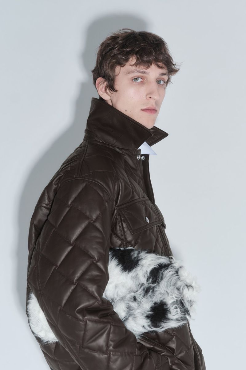 Model posing in a dark brown quilted leather jacket holding a black and white faux fur clutch against a plain white studio backdrop.