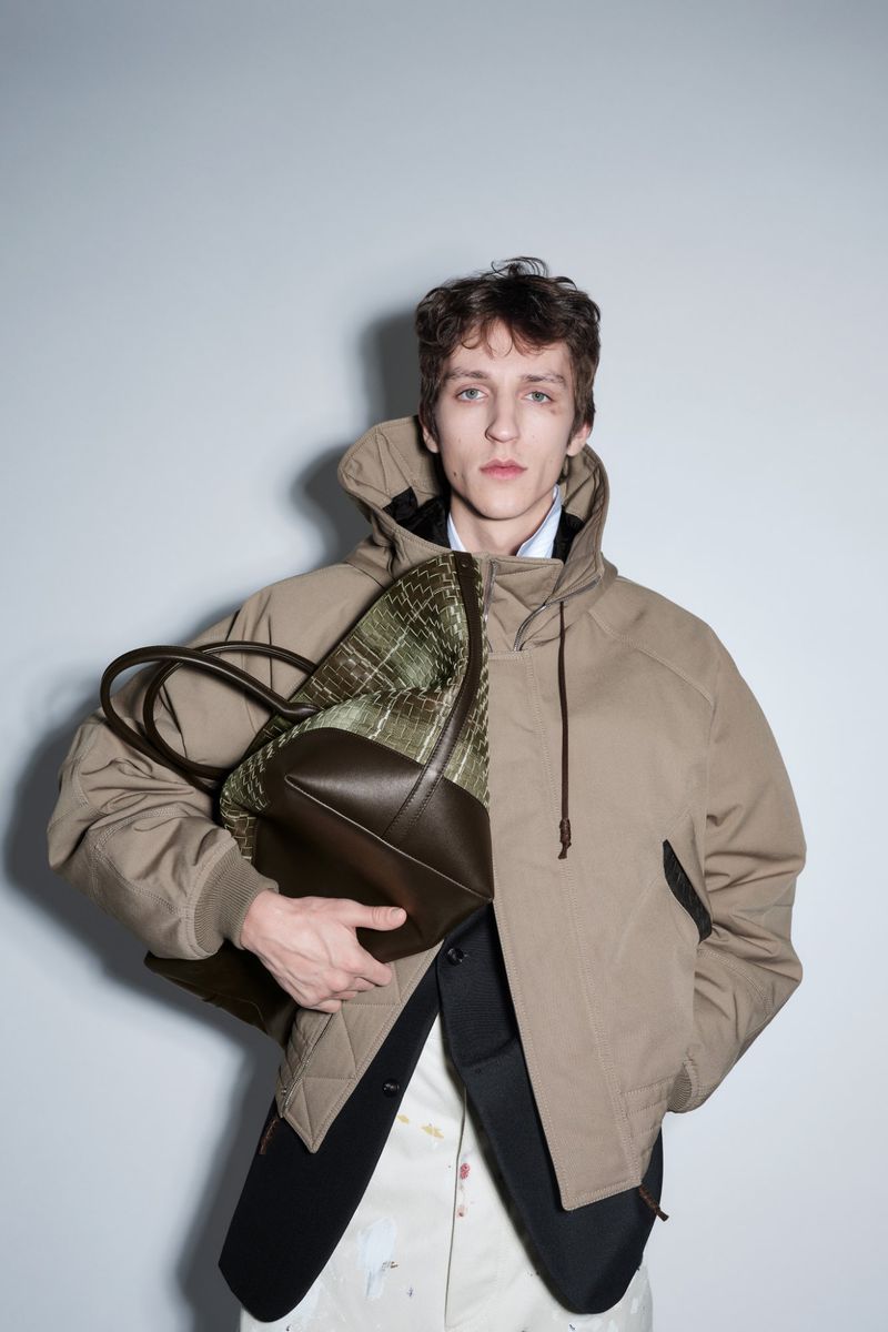Model Saul Symon wears a beige hooded jacket holding a large woven leather tote bag against a neutral studio backdrop.