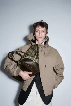 Model Saul Symon wears a beige hooded jacket holding a large woven leather tote bag against a neutral studio backdrop.