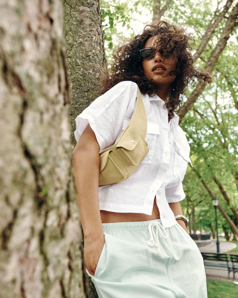 Model Nikita Wiorek leaning against a tree trunk in a white cropped shirt, mint green trousers, and a tan crossbody sling bag.