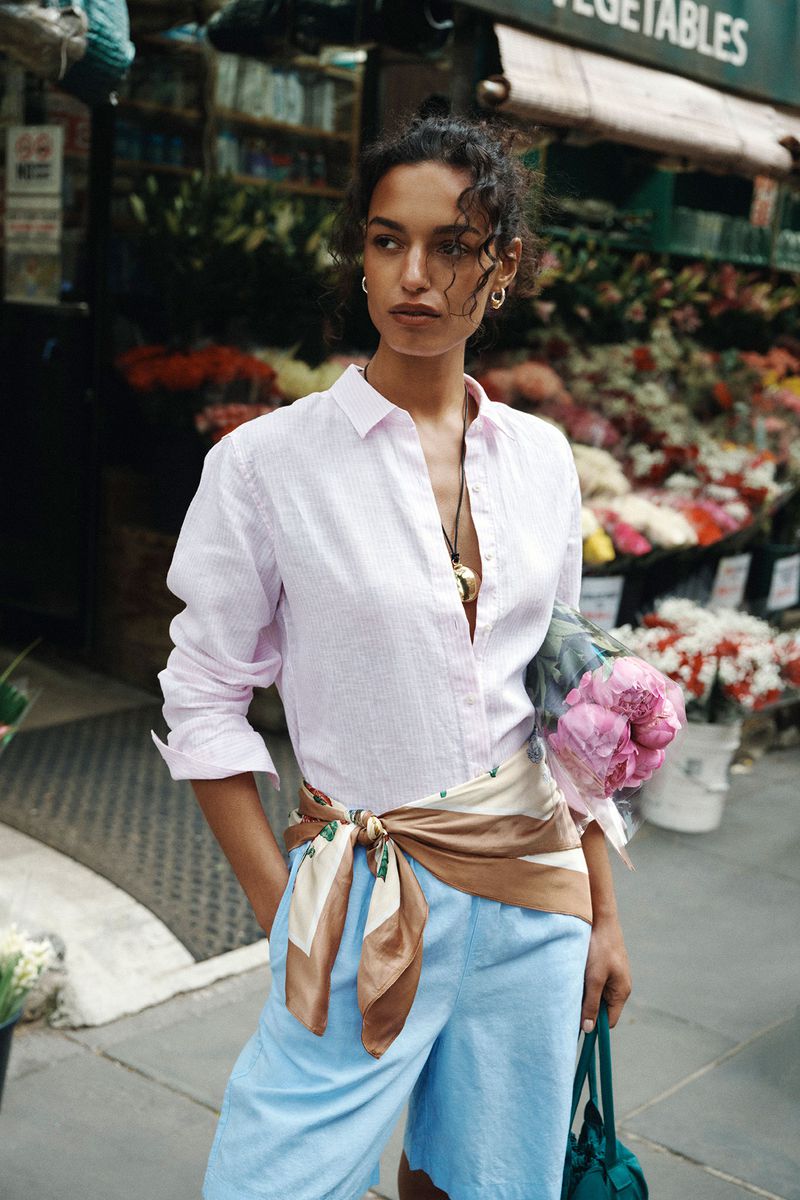Nikita Wiorek standing outside a flower stand in a light pink button-down shirt, blue shorts, and a scarf tied around her waist.