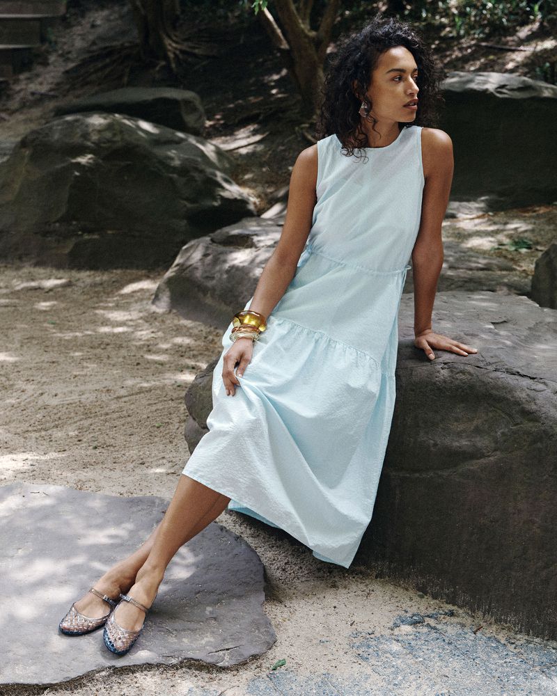 Model wearing a light blue dress seated on a large grey rock in a natural woodland setting for a Uniqlo fashion campaign.