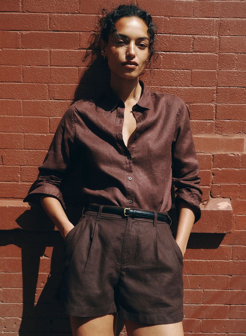 Model Nikita Wiorek in a brown linen button-down shirt and matching pleated shorts against a red brick wall in direct sunlight.
