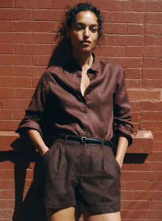 Model Nikita Wiorek in a brown linen button-down shirt and matching pleated shorts against a red brick wall in direct sunlight.