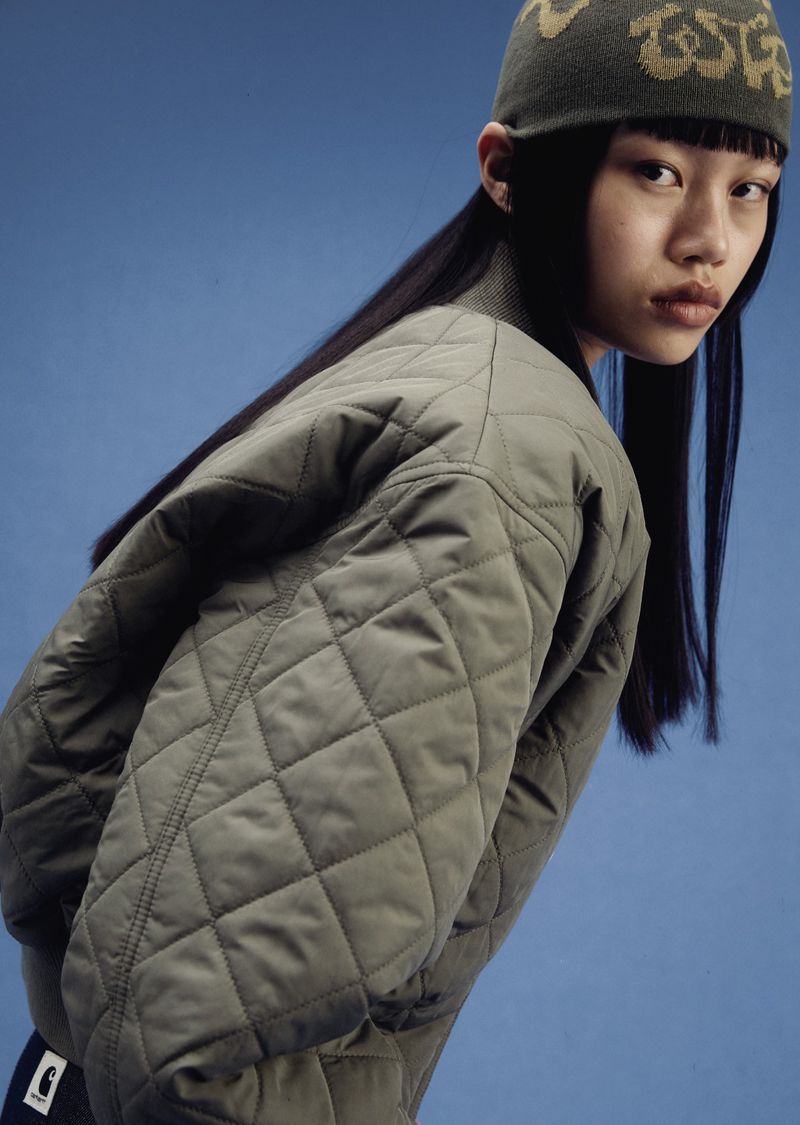 East Asian model looking over shoulder wearing an olive green diamond-quilted bomber jacket and beanie against a plain blue backdrop.
