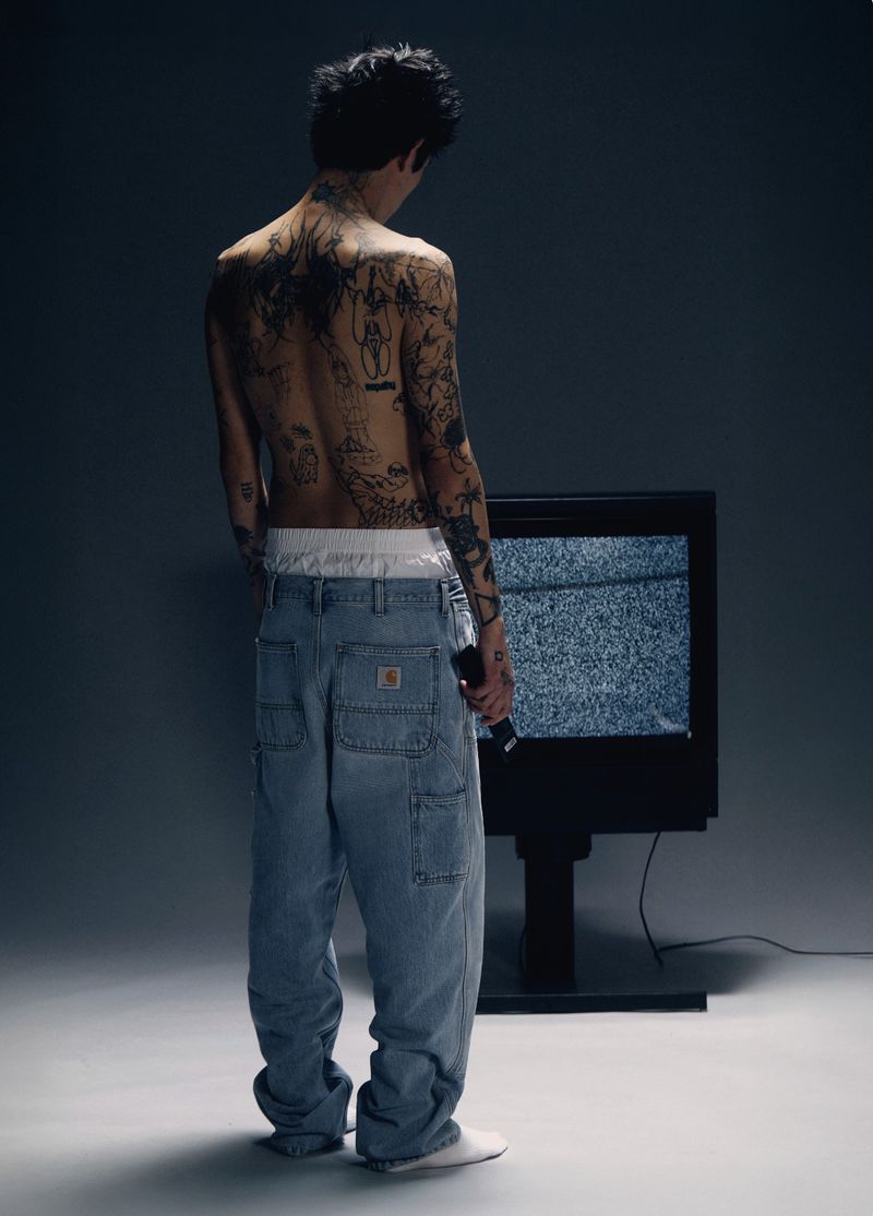 Shirtless tattooed man from behind wearing loose light blue Carhartt denim jeans in front of a static television screen.