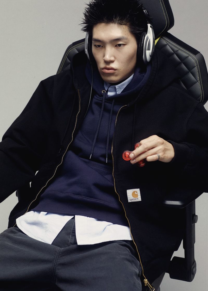 Model wearing headphones, layered Carhartt hoodie, and black zip jacket while seated in a gaming chair holding a spinner.