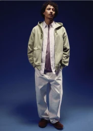 Model Jackson Hale wearing a sage zip hoodie and loose white pants against a deep blue studio backdrop for Carhartt.