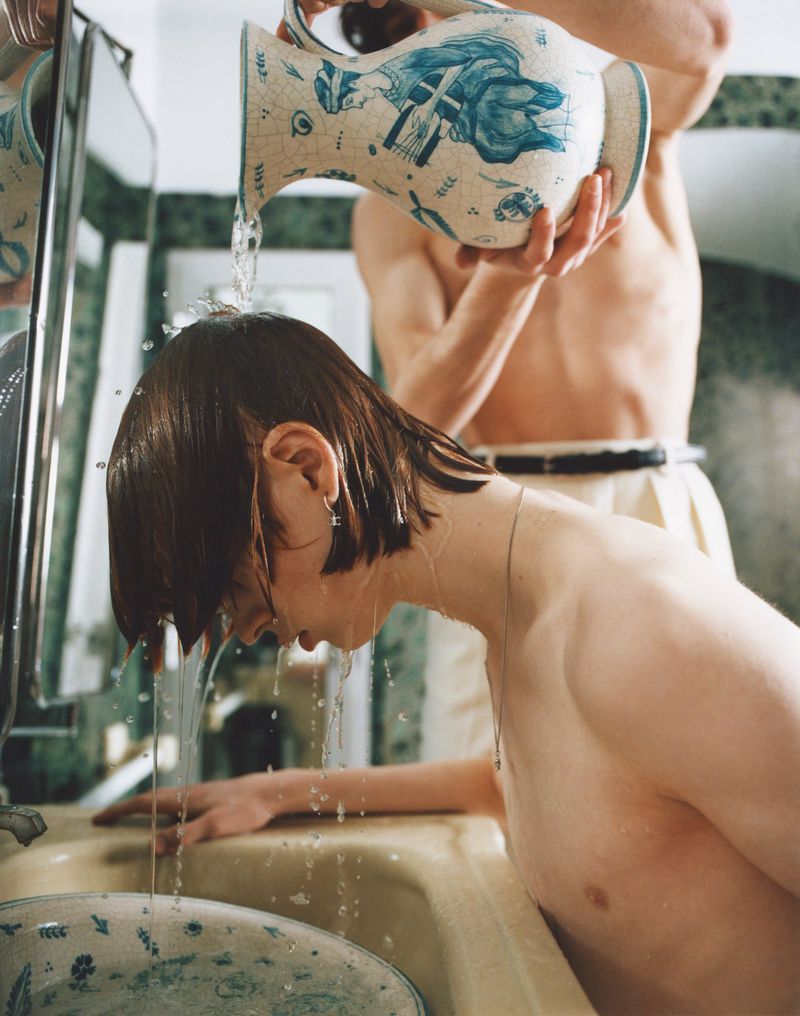 Man pours water from a patterned pitcher onto a person's head in a bathroom with green marble accents, creating a clean, refreshing feel.
