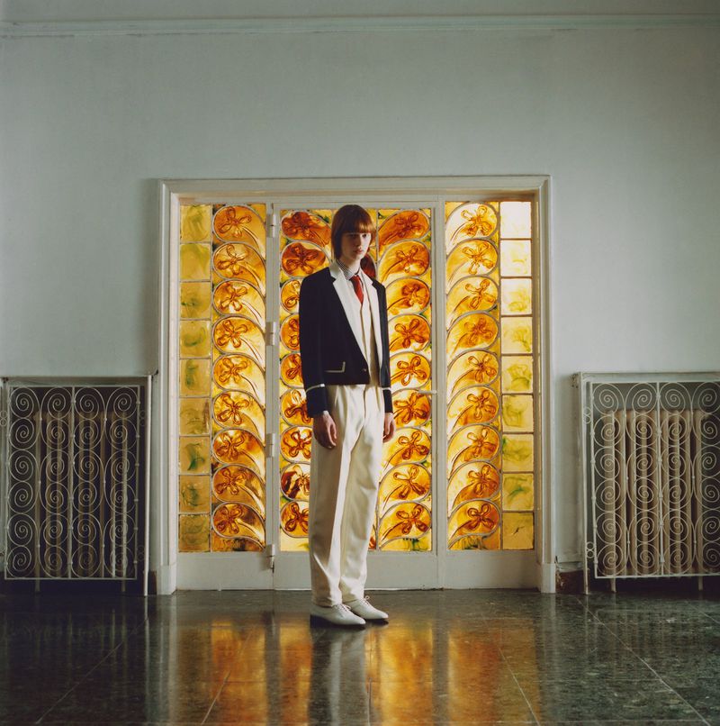Young adult stands in formal wear against an amber stained-glass window, flanked by metal radiators in an interior setting with green tile floor.