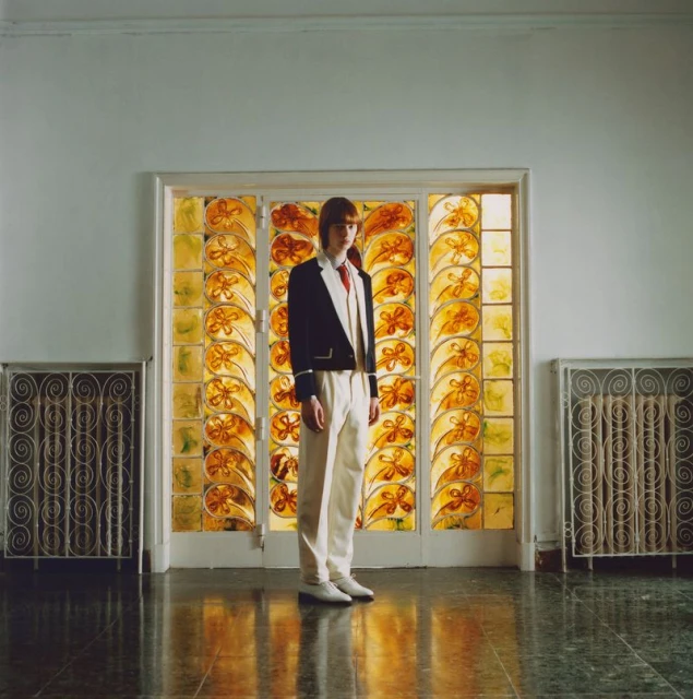 Young adult stands in formal wear against an amber stained-glass window, flanked by metal radiators in an interior setting with green tile floor.