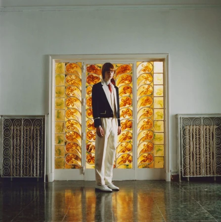 Young adult stands in formal wear against an amber stained-glass window, flanked by metal radiators in an interior setting with green tile floor.