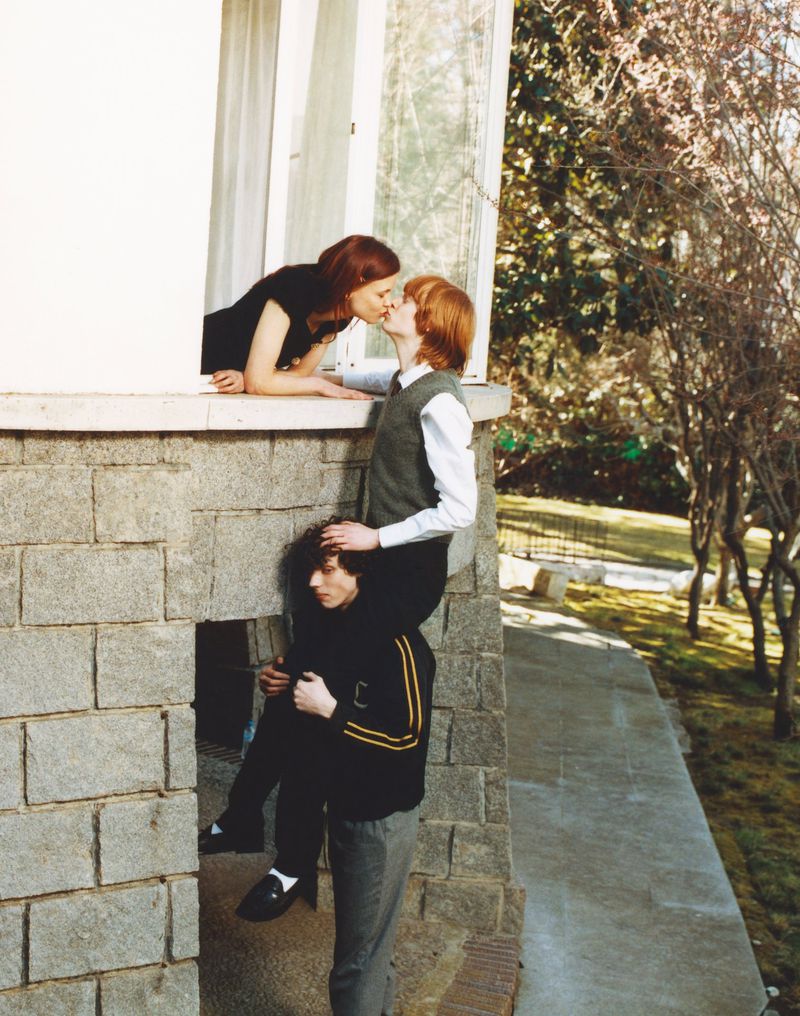 Outdoor shot features a woman and man kissing by a window, while another man carries the kissing man on his shoulders, next to a stone wall.