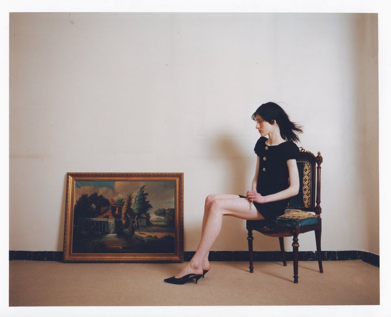 A woman sits on an ornate chair next to a framed landscape painting against a neutral wall in an indoor setting with marble baseboards.