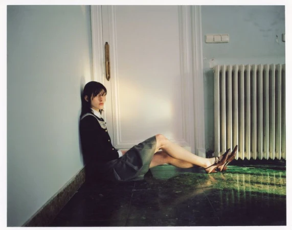 Young woman in school uniform sitting against a wall on a marble floor with radiator and door. Light from window is visible.