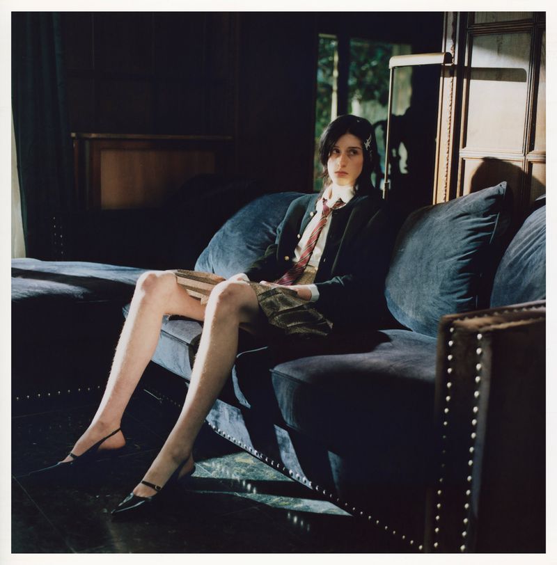 Young woman in a school uniform lounges on a dark blue tufted couch, holding a book, inside a room with dark wood paneling and window light.