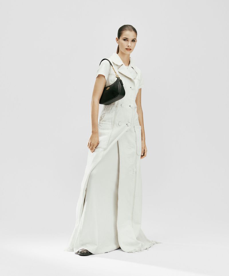 Model River Klein in a full-length white coat dress carrying a black Coach shoulder bag against a white seamless backdrop.