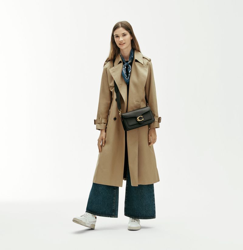 Model River Klein wears a beige trench coat, wide-leg jeans, and a black Coach crossbody bag against a white studio background.