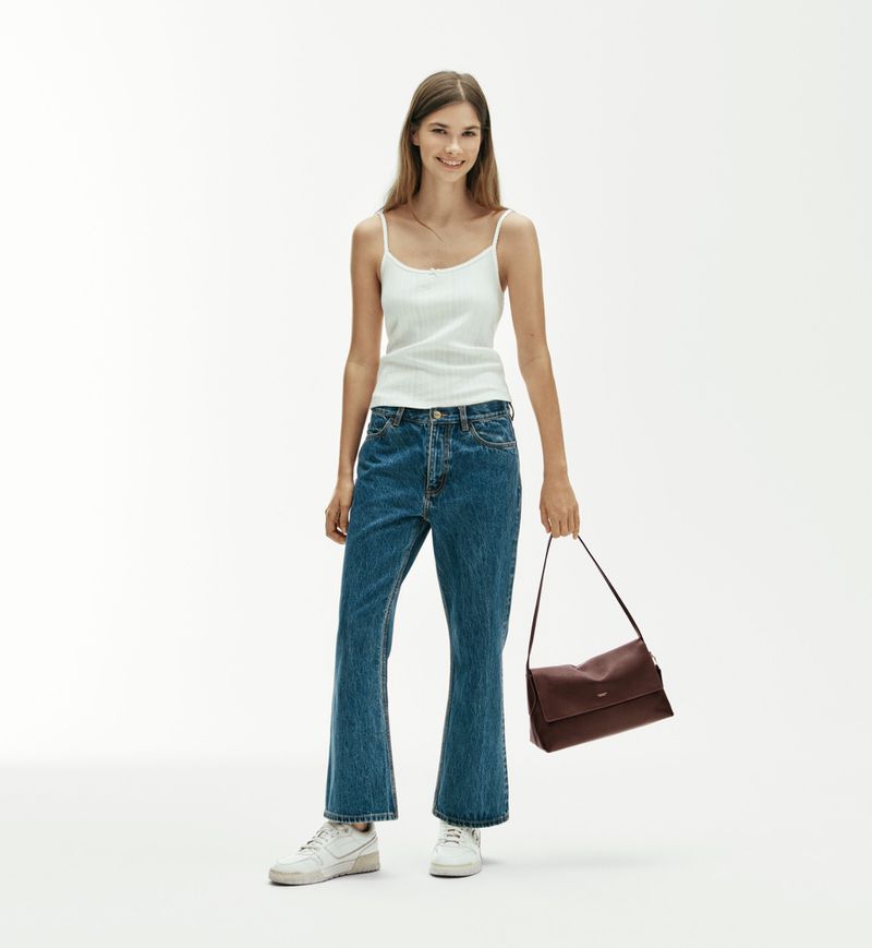 Model wearing a white tank top and blue jeans holding a dark brown handbag against a white background
