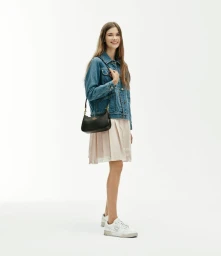 Model River Klein wearing a blue denim jacket, pale pleated skirt, and a black shoulder bag against a bright white background.